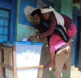 दुर्गाथली ५ महादेव प्रावि मतदान केन्द्रमा बोकेर एक वृद्धा महिलालाई मतदान गराइँदै